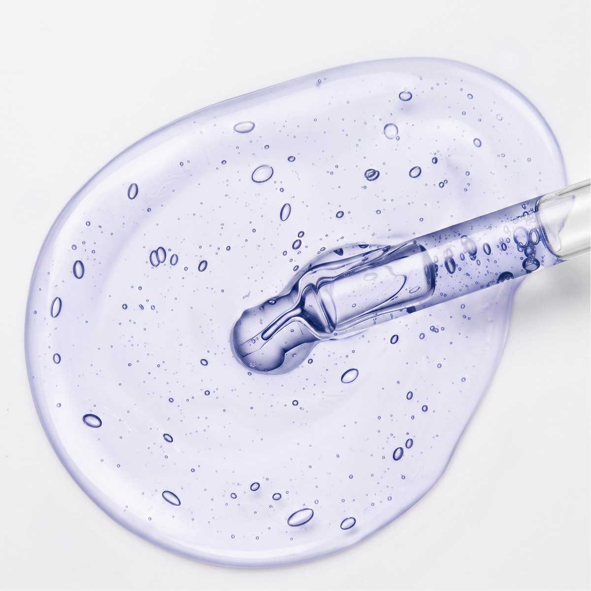 A close-up of a copper peptide translucent lavender-blue cosmetic gel or serum droplet spread on a white surface, with a glass pipette dipping into the center.