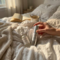 Hand holding a skincare product on a bed with books and a cup of coffee.